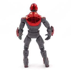 Op de afbeelding: Een grijs actiefiguur met rode accenten, van achteren gezien. De figuur heeft een rode helm en borstplaat met een metallic glans. Rode details zijn ook aanwezig op de schouders, onderarmen en onderbenen. De figuur staat tegen een witte achtergrond.