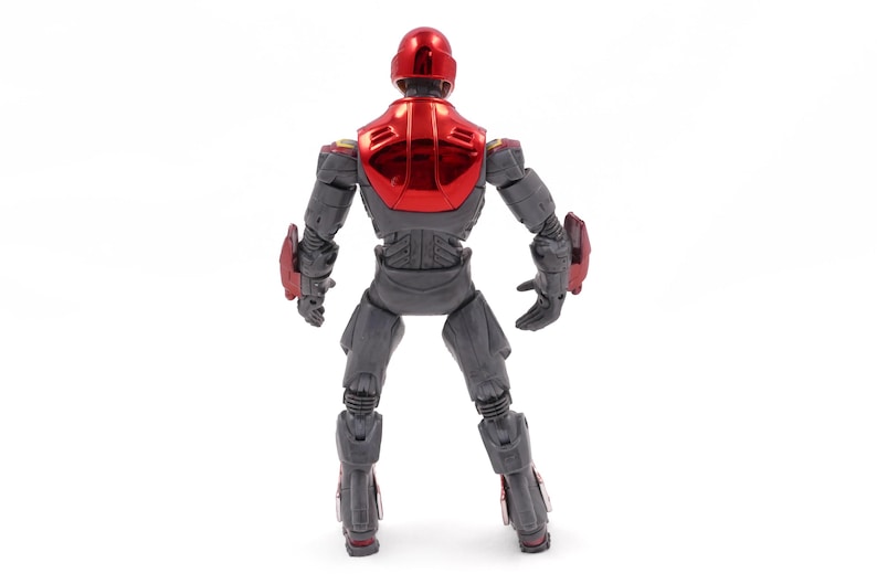 Op de afbeelding: Een grijs actiefiguur met rode accenten, van achteren gezien. De figuur heeft een rode helm en borstplaat met een metallic glans. Rode details zijn ook aanwezig op de schouders, onderarmen en onderbenen. De figuur staat tegen een witte achtergrond.