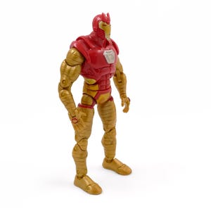 May include: A red and gold action figure of a superhero, standing upright. The figure has a red helmet and chest, with gold-colored arms, legs, and feet. A silver emblem is on the chest. The figure is posed against a white background.
