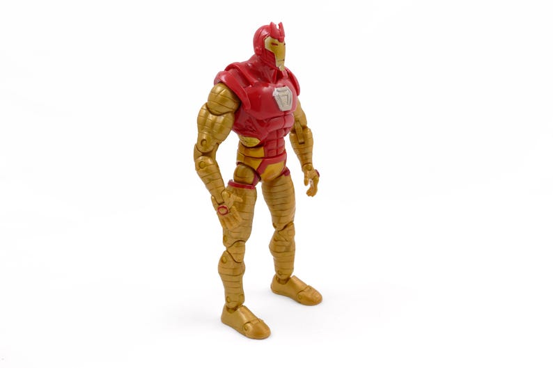 May include: A red and gold action figure of a superhero, standing upright. The figure has a red helmet and chest, with gold-colored arms, legs, and feet. A silver emblem is on the chest. The figure is posed against a white background.