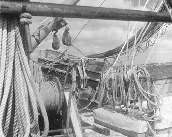 May include: A black and white photo of a boat's deck with a large spool of rope, a winch, and various ropes and pulleys.