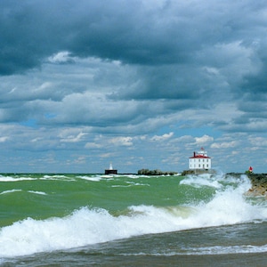 Könnte beinhalten: Ein weißer Leuchtturm mit rotem Dach steht auf einem felsigen Vorsprung inmitten eines unruhigen, grünen Sees. Der Himmel ist bewölkt mit einer Mischung aus dunkelgrauen und weißen Wolken.