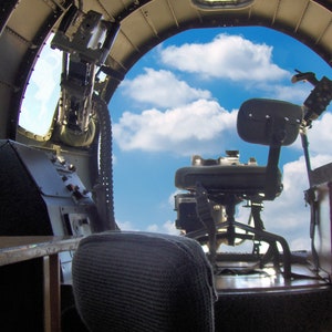 Puede incluir: El interior de una cabina de avión vintage con un asiento de artillero y una torreta de ametralladora. La vista desde la ventana de la cabina muestra un cielo azul con nubes blancas.