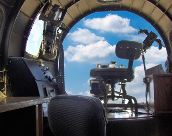 May include: A vintage airplane cockpit interior with a gunner's seat and a machine gun turret. The view from the cockpit window shows a blue sky with white clouds.