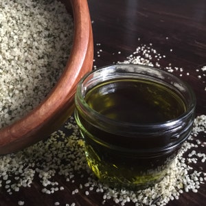 May include: A glass jar filled with dark green liquid, probably hemp seed oil, sits on a wooden table surrounded by hemp seeds.