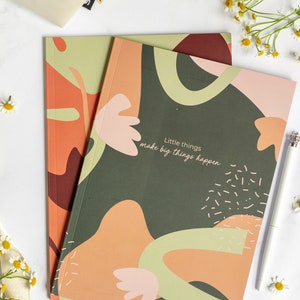 Könnte beinhalten: Zwei Notizbücher mit abstrakten Designs in Grün-, Orange- und Rosatönen. Das obere Notizbuch trägt den Text "Little things make big things happen" und das untere Notizbuch hat das Wort "SEWK" unten rechts gedruckt.