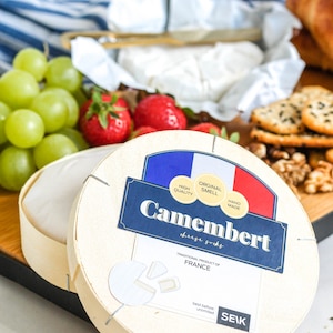 Könnte beinhalten: Eine runde Holzkiste mit einem weißen Etikett, auf dem "Camembert cheese greks" und "TRADITIONAL PRODUCT OF FRANCE" steht. Das Etikett hat auch ein französisches Flaggen-Design in Blau, Weiß und Rot mit den Worten "ORIGINAL SMELL", "HIGH QUALITY" und "HAND MADE".
