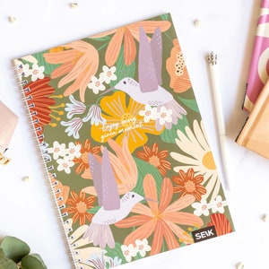 Op de afbeelding: Een groene spiraalgebonden notitieboek met een bloemenontwerp met kolibries en de tekst "Enjoy every given moment". Het notitieboek heeft een bruine achtergrond met roze, oranje, gele en witte bloemen.