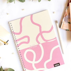 May include: A pink and cream spiral bound notebook with white abstract lines and the text "BE YOU NOT THEM" in pink. The notebook has a black logo that says "SEK" in the bottom right corner.