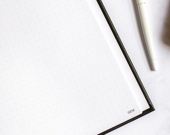 May include: A black and white dotted notebook with a black cover and the word "SEIK" printed on the bottom right corner. A white pen is lying on the notebook.