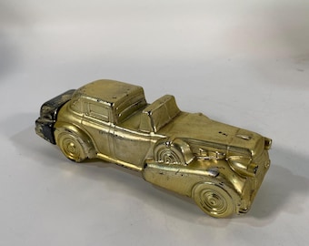 May include: A vintage-style toy car with a metallic gold finish and a black rear trunk section. The model features a long hood, prominent wheel arches, and a cabin with an open top. The gold paint shows signs of wear and aging throughout.