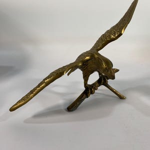 May include: A brass sculpture of a bird with outstretched wings, perched on a branch. The bird is detailed with textured feathers and a sharp beak. The sculpture has a golden hue and is set against a white background.