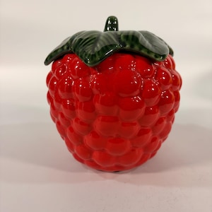 May include: A ceramic lidded jar shaped like a bright red raspberry with a textured, bumpy surface. The lid features a dark green stem and leaf design that sits atop the rounded red body, creating a realistic fruit-inspired container.