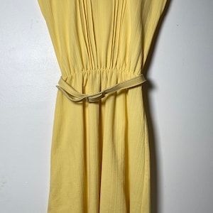 Puede incluir: Un vestido amarillo sin mangas con cuello en V y cinturón. El vestido tiene un corpiño plisado y una cintura fruncida.