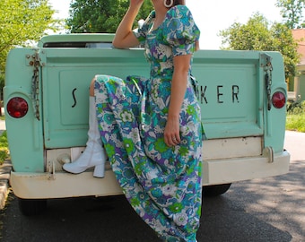 May include: A woman wearing a blue and green floral maxi dress with white boots, standing on the back of a light blue pickup truck. The truck has the word "SOKER" written on the back.