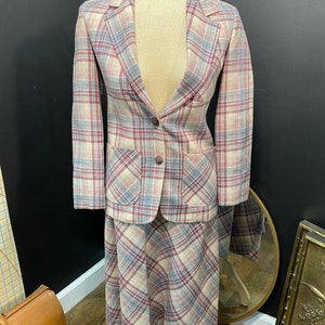 May include: A vintage plaid skirt suit in beige, red, and blue. The blazer has two front pockets and a single button closure. The skirt is a midi length. The suit is displayed on a mannequin.