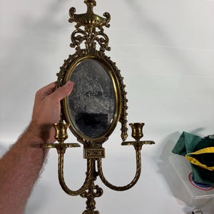 May include: A gold-toned metal wall sconce featuring an oval mirror at the center, flanked by two candle holders on curved arms. The frame is decorated with ornate scrollwork and a finial at the top and bottom, creating a symmetrical design.