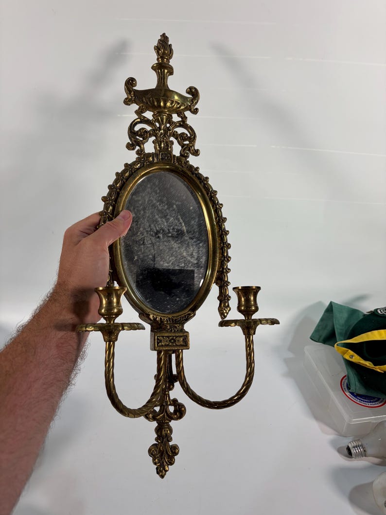 May include: A gold-toned metal wall sconce featuring an oval mirror at the center, flanked by two candle holders on curved arms. The frame is decorated with ornate scrollwork and a finial at the top and bottom, creating a symmetrical design.