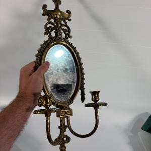 May include: An ornate brass wall sconce featuring a central oval mirror framed by intricate scrollwork and decorative finials. The fixture includes two curved arms extending outward, each topped with a circular candle cup holder for taper candles.