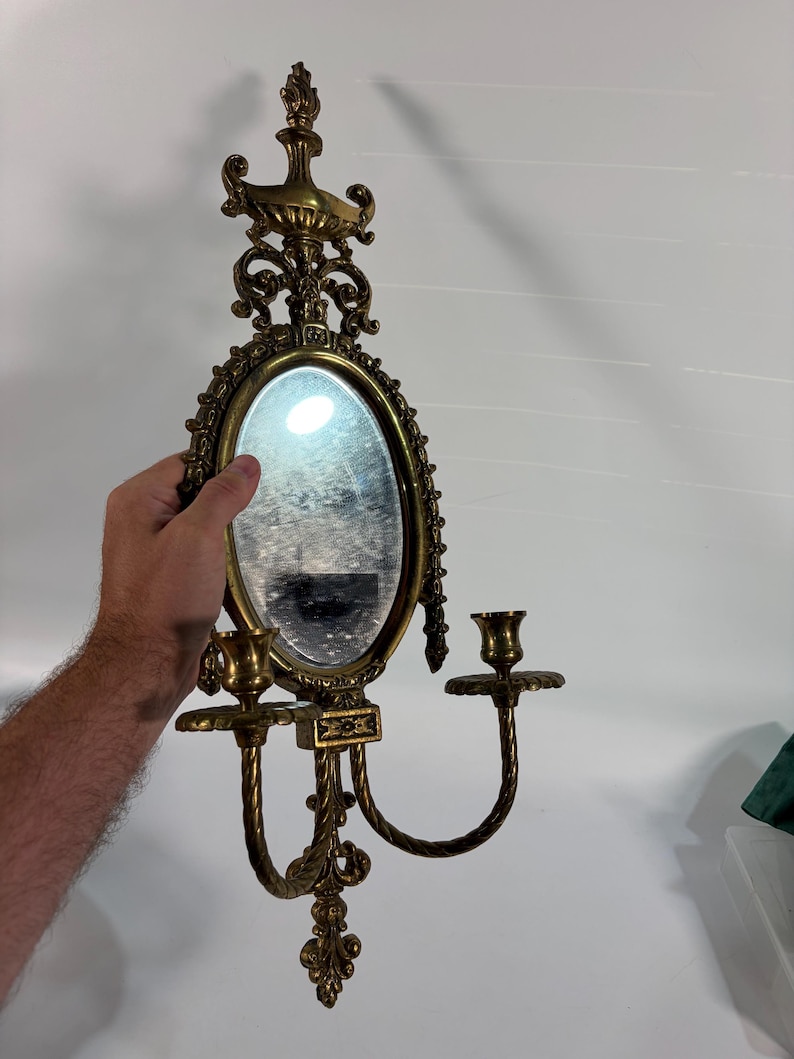May include: An ornate brass wall sconce featuring a central oval mirror framed by intricate scrollwork and decorative finials. The fixture includes two curved arms extending outward, each topped with a circular candle cup holder for taper candles.
