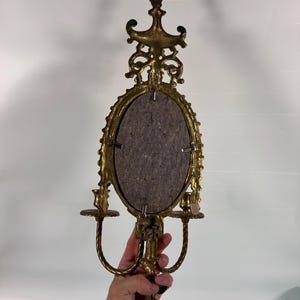 May include: A gold-toned metal wall sconce featuring an oval center frame with a dark gray backing, ornate scrollwork detailing at the top and bottom, and two candle holders extending from the sides with twisted metal support arms.