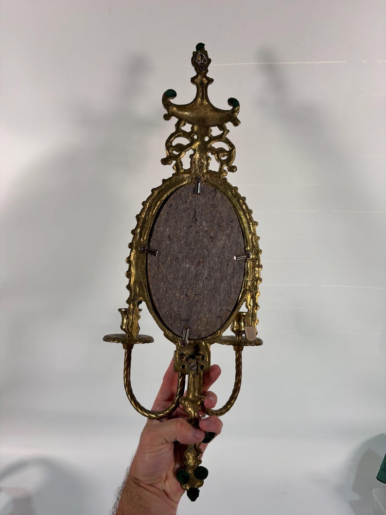 May include: A gold-toned metal wall sconce featuring an oval center frame with a dark gray backing, ornate scrollwork detailing at the top and bottom, and two candle holders extending from the sides with twisted metal support arms.