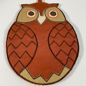 May include: A decorative owl-shaped pot holder in shades of orange, tan, and brown. The pot holder features a stitched design with a loop for hanging. The owl's eyes are brown with orange accents, and the wings have a zig-zag pattern.