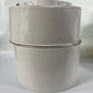 May include: A two-piece, off-white, cylindrical hat box with a shiny, glossy finish. The top section has a small handle. The two sections are joined by a silver zipper. The box is made of a durable material and is designed to protect hats.