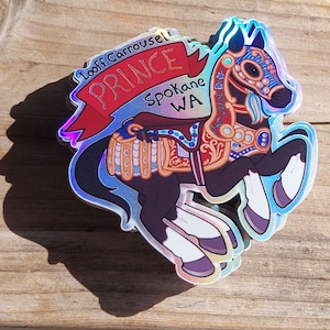 May include: Holographic sticker featuring a carousel horse named "Prince" with ornate detailing in red, gold, and blue. The sticker includes text that reads "Looff Carrousel" and "Spokane WA". The horse has a black body and white legs.