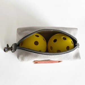Puede incluir: Una bolsa de lona gris con cremallera y una lengüeta de cuero marrón. La bolsa está abierta y contiene dos pelotas de pickleball amarillas.