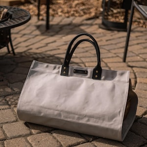 May include: A light grey tote bag with black leather handles and accents. The bag has a rectangular shape and is on a brick patio. The handles are attached with gold-coloured rivets.