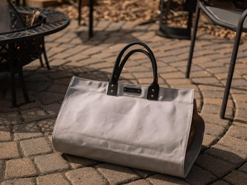 May include: A light grey tote bag with black leather handles and accents. The bag has a rectangular shape and is on a brick patio. The handles are attached with gold-coloured rivets.