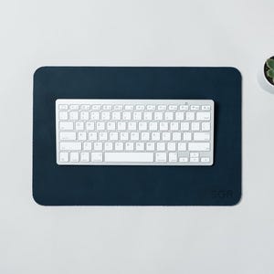 May include: A navy blue rectangular desk mat with rounded corners, a white keyboard, and a small potted succulent. The mat has the initials "SGR" in the bottom right corner. The keyboard is centered on the mat.