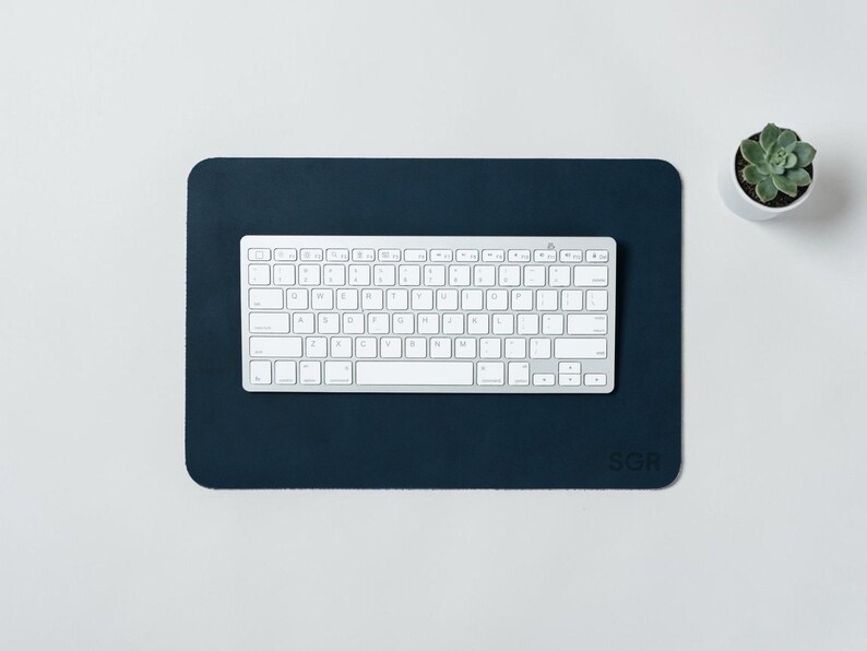 May include: A navy blue rectangular desk mat with rounded corners, a white keyboard, and a small potted succulent. The mat has the initials "SGR" in the bottom right corner. The keyboard is centered on the mat.
