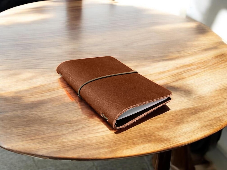 Puede incluir: Un diario de cuero marr&oacute;n con un cierre el&aacute;stico a juego descansa sobre una mesa de madera. El diario rectangular parece estar lleno de p&aacute;ginas blancas. La mesa tiene un acabado de veta de madera clara.