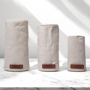 May include: Three cylindrical covers in varying heights made of light beige fabric. Each cover features a rectangular brown leather patch at the base, secured with two gold-toned metal rivets, and embossed with the letters KP in the center.