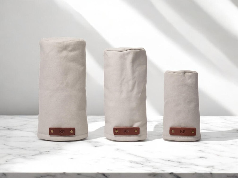 May include: Three cylindrical covers in varying heights made of light beige fabric. Each cover features a rectangular brown leather patch at the base, secured with two gold-toned metal rivets, and embossed with the letters KP in the center.