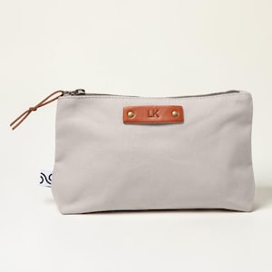 May include: A light gray canvas pouch with a brown leather tag featuring the initials "LK" and secured with two brass rivets. The pouch has a brown zipper and a matching leather pull. A small tag with a black and white design is attached to the side.