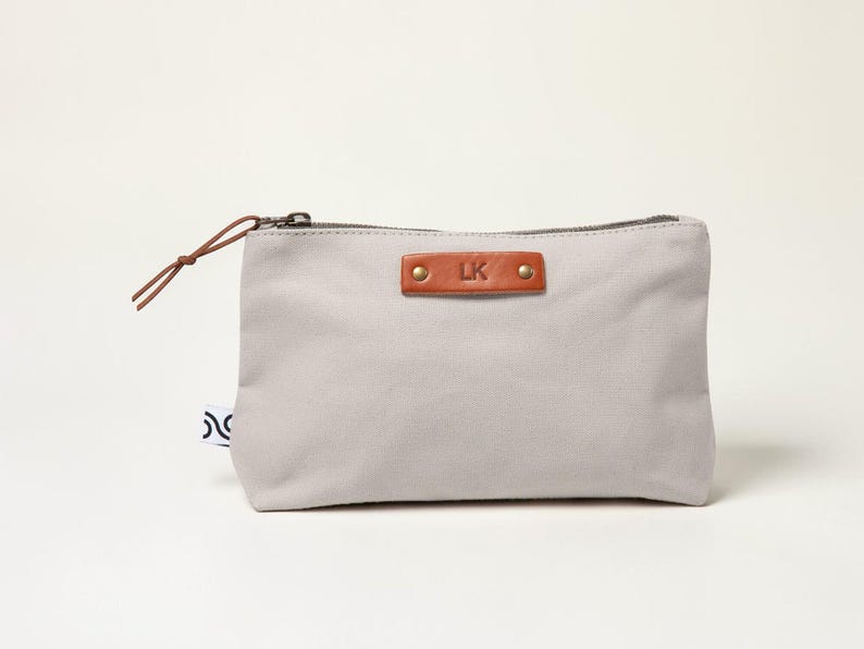 May include: A light gray canvas pouch with a brown leather tag featuring the initials "LK" and secured with two brass rivets. The pouch has a brown zipper and a matching leather pull. A small tag with a black and white design is attached to the side.