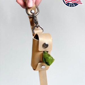 May include: A light tan leather waste bag dispenser attached to a matching leather leash with a silver metal swivel snap hook and rivets. The cylindrical holder features a central opening revealing a green plastic waste bag inside.
