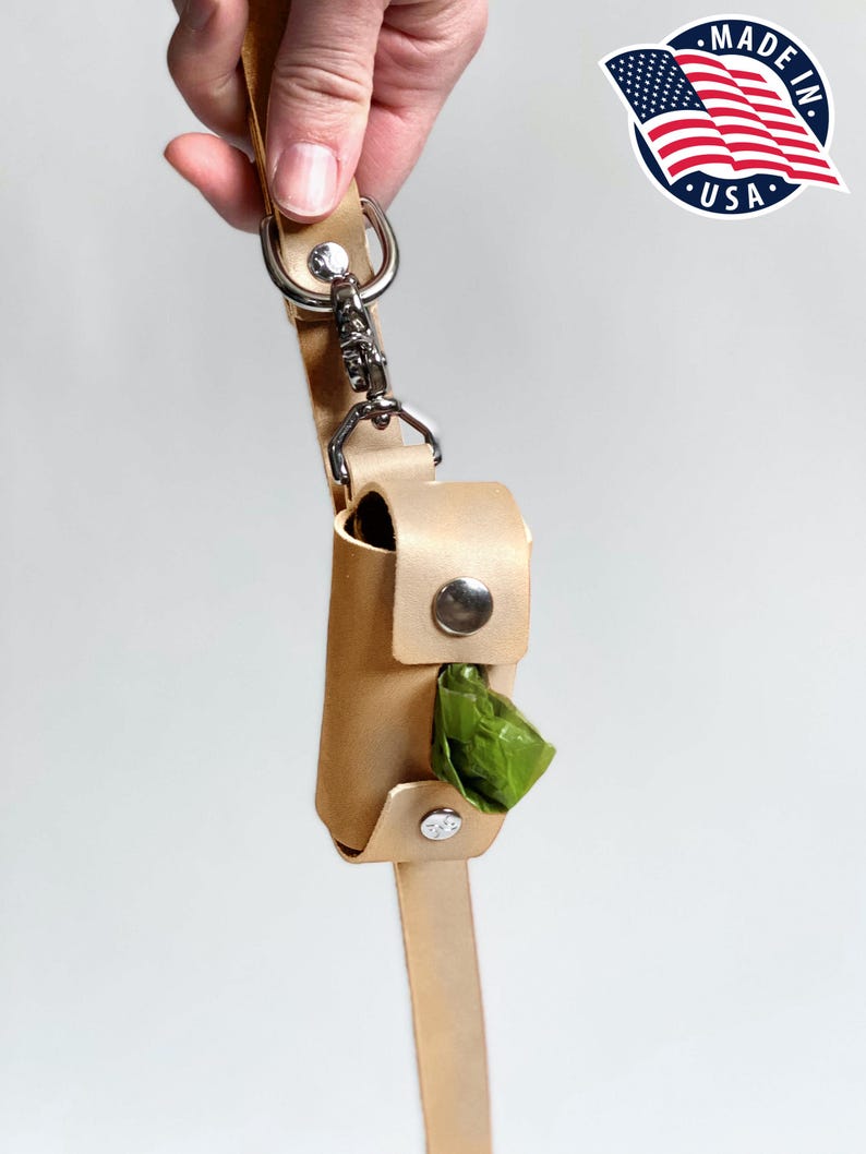 May include: A light tan leather waste bag dispenser attached to a matching leather leash with a silver metal swivel snap hook and rivets. The cylindrical holder features a central opening revealing a green plastic waste bag inside.