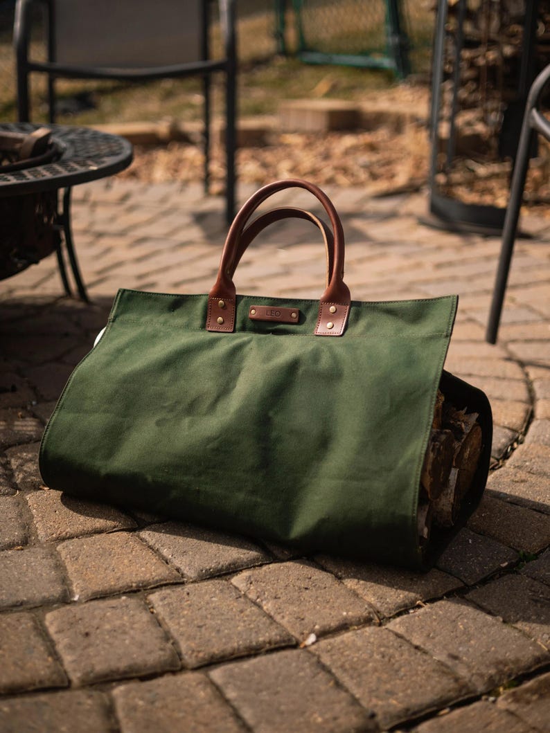May include: A dark green canvas log carrier with brown leather handles and accents. The carrier is filled with firewood and rests on a brick patio. The word "LEO" is embossed on a small leather tag.