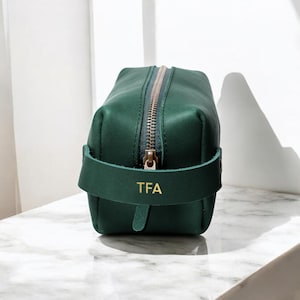 Puede incluir: Un neceser de cuero verde oscuro con cremallera dorada y asa. El asa tiene las iniciales doradas "TFA". La bolsa rectangular está sobre una superficie de mármol blanco. Un accesorio elegante.