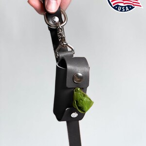 May include: A black leather waste bag dispenser attached to a matching black leather leash with a silver metal swivel snap hook and D-ring. The dispenser features a silver snap button closure and contains a single green plastic waste bag.