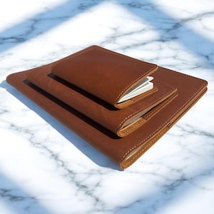 May include: Three brown leather notebooks of different sizes are stacked on a white marble surface. The notebooks have rounded corners and visible stitching. The smallest notebook is open, revealing white pages.