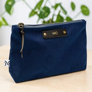 May include: A navy blue zippered pouch with a leather tag displaying the initials "HC". The pouch has a bronze-colored zipper and a leather pull. A small white tag with a black design is attached to the side. The pouch is sitting on a light-colored wooden surface.