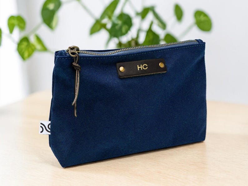 May include: A navy blue zippered pouch with a leather tag displaying the initials "HC". The pouch has a bronze-colored zipper and a leather pull. A small white tag with a black design is attached to the side. The pouch is sitting on a light-colored wooden surface.