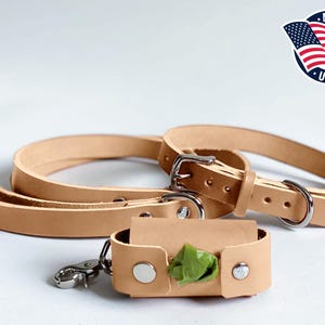 May include: A natural tan leather dog leash and collar set with silver-toned metal hardware, including a buckle and D-ring. A matching tan leather waste bag dispenser is attached with a silver metal swivel clip, containing a green plastic bag.