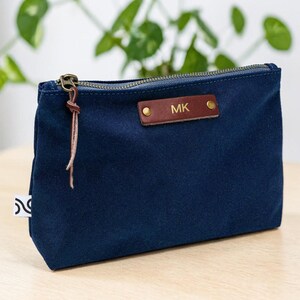 May include: A navy blue canvas pouch with a brass zipper and a leather tag. The tag is embossed with the initials "MK". A leather pull is attached to the zipper. The pouch is sitting on a light wood surface.