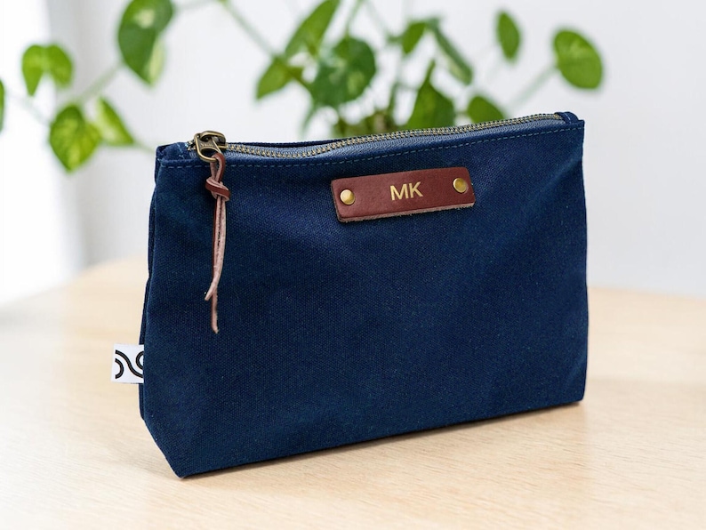May include: A navy blue canvas pouch with a brass zipper and a leather tag. The tag is embossed with the initials "MK". A leather pull is attached to the zipper. The pouch is sitting on a light wood surface.